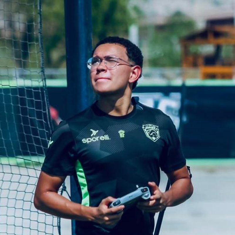 Higor Santos, professor do curso, durante um treino, de uniforme técnico e com o controle de uma câmera na mão.